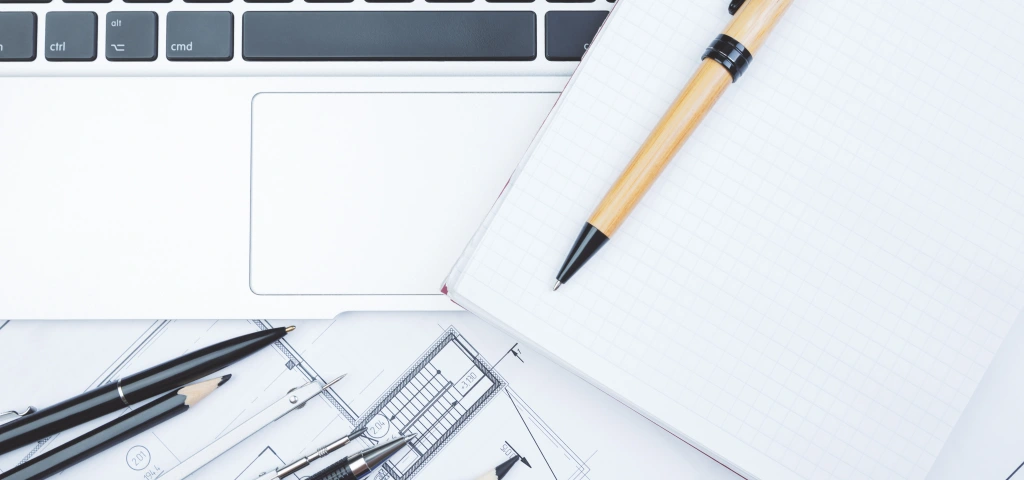 Cuaderno, bolígrafo y planos junto a un portátil para preparar el briefing de una página web