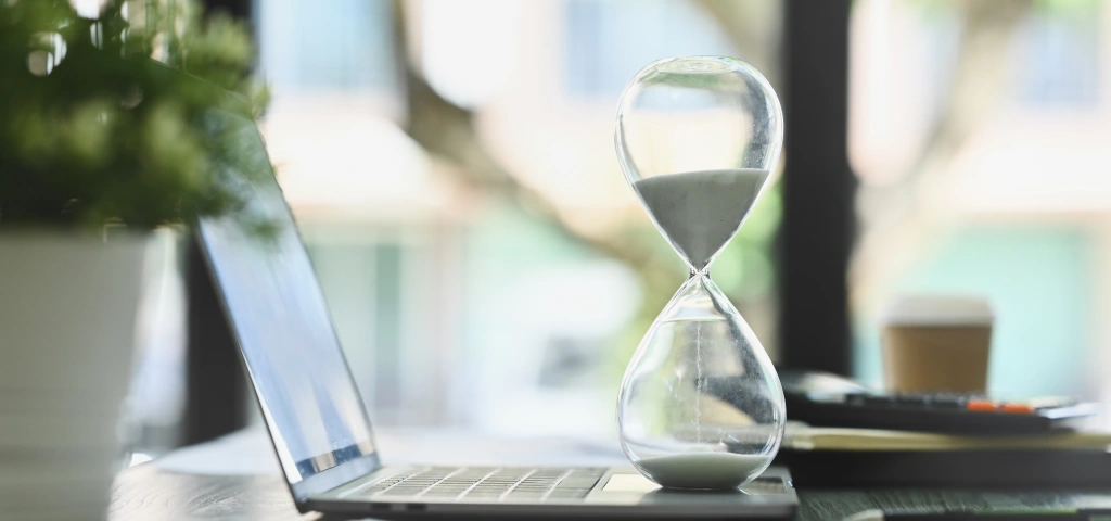 Reloj de arena sobre la mesa junto a un portátil, para ilustrar cuánto se tarda en hacer una página web